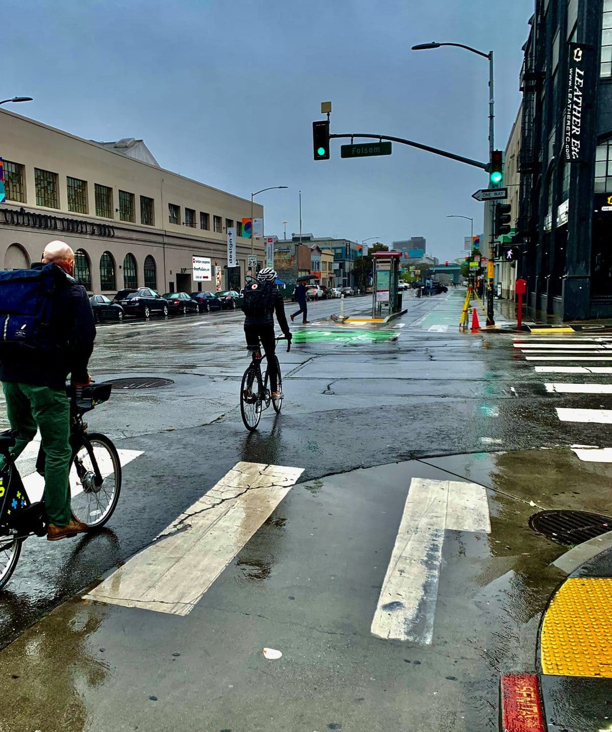 Two people riding bicycles in a protected bike lane with a cloudy gloomy sky and water on the road around them like it has just rained.