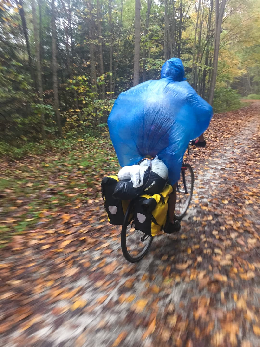 A person on a bicycle with a rain poncho billowing in the wind. The rear rack has 2 large panniers and some other things strapped on with a bungie cord.