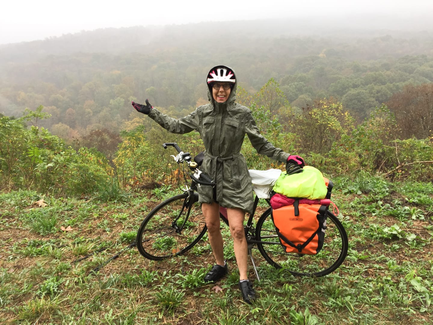 A person in a rain jacket standing next to a bicycle with all the touring gear and panniers. A beautiful foggy tree filled landscape is in the background.