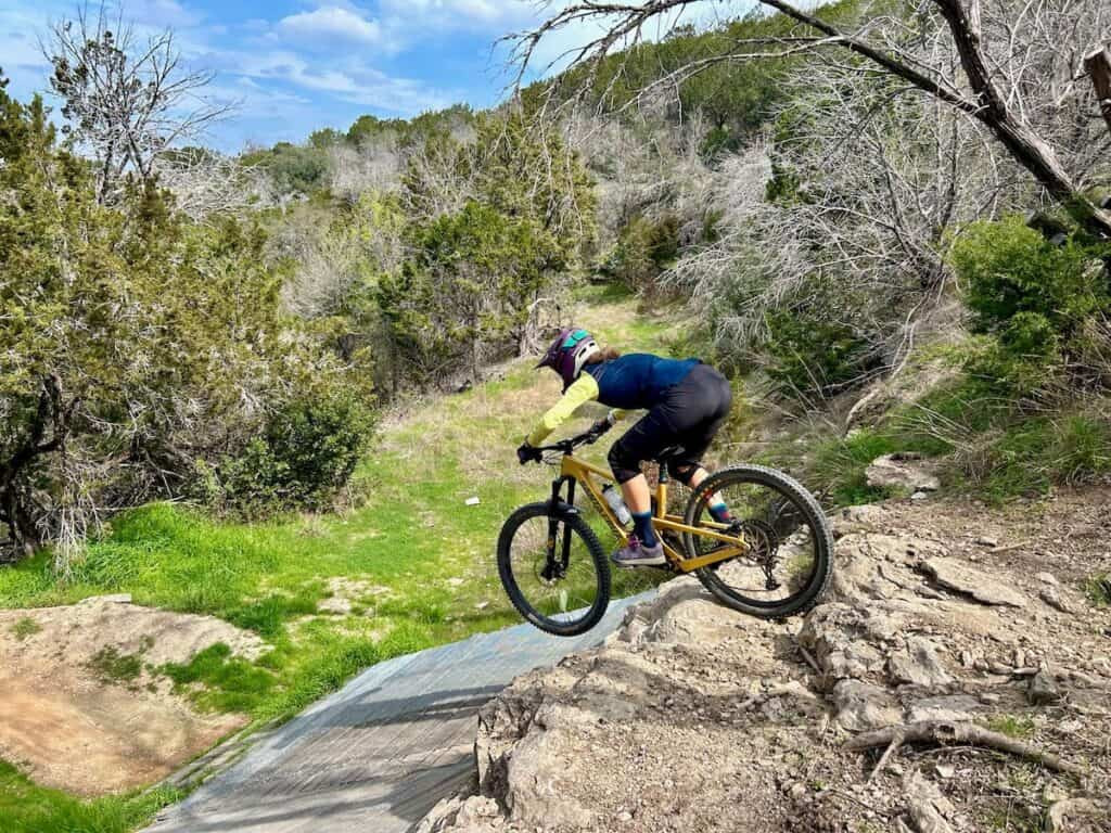 A mountain biker confidently rides off a rock ledge onto a wooden ramp at Spider Mountain Bike Park in Texas