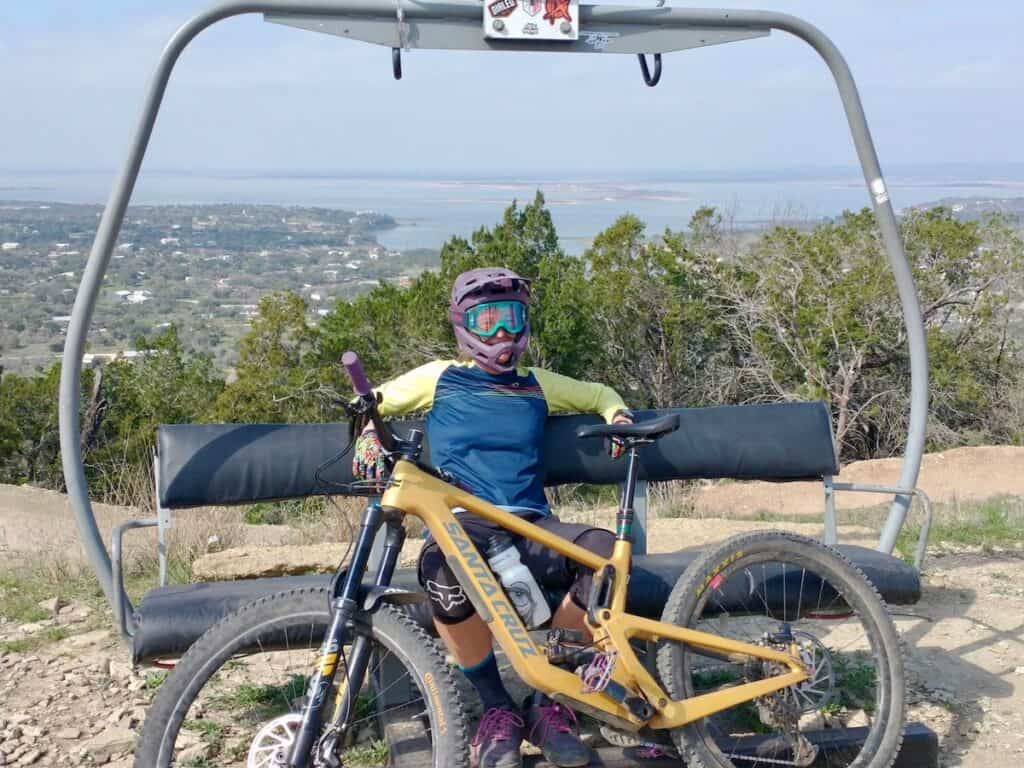 A woman sits on a chairlift with her mountain bike, enjoying the scenic views of Lake Buchanan at Spider Mountain Bike Park in Texas