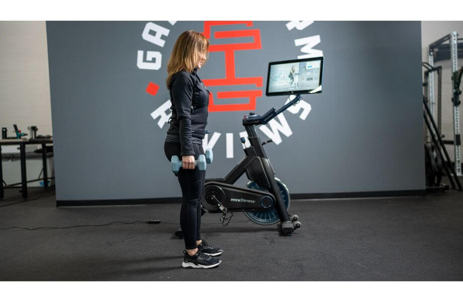 Woman standing with dumbbells next to the MYX II bike, highlighting its compact design and versatility for cross-training.
